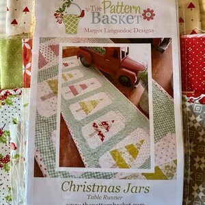 以下が含まれることがあります： クリスマスジャーのデザインが施された白と緑の布製のテーブルランナー。パターンには赤いトラックと「クリスマスジャー」と「テーブルランナー」の文字が書かれています。このパターンはザ・パターンバスケットから入手できます。