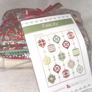 以下が含まれることがあります： 赤いリボンで結ばれた透明なビニール袋には、布の束と「Felicity」というタイトルのパターンが入っています。パターンは、さまざまな色のクリスマスオーナメントのデザインが特徴です。布の束は、赤、緑、白のパターンを持っています。