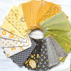 May include: A collection of fabric squares arranged in a circular fan pattern, featuring a variety of patterns including floral prints, geometric grids, and polka dots in a color palette of yellow, olive green, gray, and white.