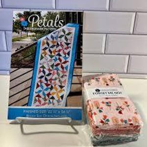 May include: A sewing pattern book titled "Petals" with a finished table runner size of 56 cm x 137 cm. Beside it is a stack of fabric squares with a floral pattern. The background is a white brick wall.