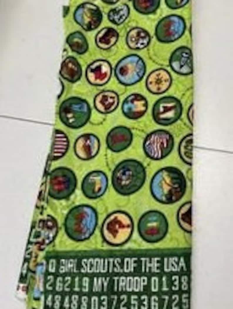 Puede incluir: Tela verde con un patr&oacute;n repetido de insignias de Girl Scouts en c&iacute;rculos blancos y marrones. La tela tiene el texto "Girl Scouts of the USA" y un n&uacute;mero de tropa.