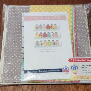 Może przedstawiać: Zestaw do mini kołdry Cherry Jam od Fat Quarter Shop. Zestaw zawiera różne tkaniny w kolorze czerwonym, żółtym, zielonym i niebieskim w kropki. Gotowa kołdra ma rozmiar 54,6 cm x 44,5 cm.