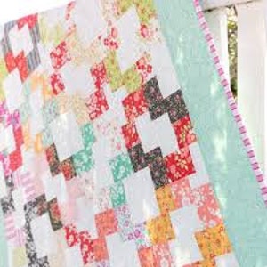 Puede incluir: Una colcha de retazos con fondo blanco y cuadrados coloridos en varios patrones florales. La colcha está bordeada con una tela azul claro y un ribete rosa y blanco.