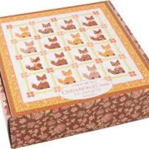 May include: A square box with a floral pattern on the sides and a decorative top featuring a grid of fox illustrations in various colors. The top has a border of orange and gold, with the text "Cinnamon & Co." on an oval label.