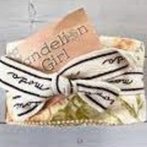 May include: A small, white gift box with a floral pattern and a white ribbon with black stripes. The box is tied with a bow and has a small card that says "Dandelion Girl" on it.