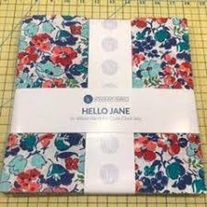 May include: A stack of floral fabric squares featuring red, teal, and navy flowers on a white background. The fabric is wrapped with a white band that reads "HELLO JANE" and "10 inch Layer Cake Box". The fabric is on a yellow cutting mat.