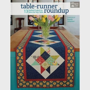 May include: A quilted table runner with a blue background and a red border. The runner features a starburst design and is decorated with yellow flowers. The book cover reads "table-runner roundup" and "13 Quilted Projects to Spice Up Your Table."