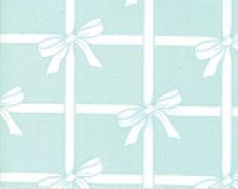 May include: A repeating pattern of light blue squares with white bows and vertical and horizontal white stripes. The fabric has a dotted texture. A ruler is at the bottom of the image, with the word "moda" printed on it.