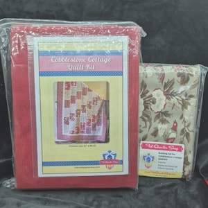 Op de afbeelding: Een rode Cobblestone Cottage Quilt Kit in een plastic verpakking, met een afgewerkte maat van 157,5 cm x 203,8 cm. Daarnaast een stoffen bundel met een bloemen- en vogelpatroon. De kit bevat een backing set.