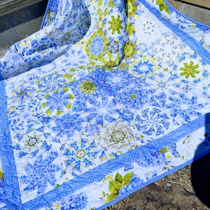 Puede incluir: Un edredón blanco con un estampado floral azul y un borde azul. El edredón presenta un diseño repetido de hortensias azules y hojas verdes. El edredón está hecho de una tela suave y acolchada.