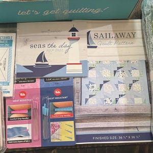 Könnte beinhalten: Ein Quilt-Set bestehend aus einer Schachtel mit der Aufschrift let's get quilting, einem Half Rectangle Trimmer, einem Seas the Day July 2022 Heft, einem Sailaway Quilt-Schnittmuster, hitzebeständigen Stecknadeln und einer Rolle blau-weißer Blumenstoffstreifen.