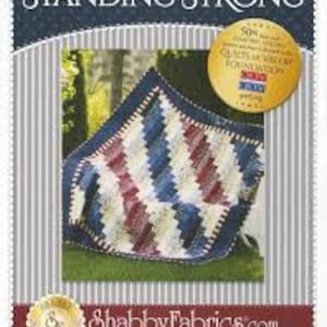 May include: A red, white and blue quilt with a geometric pattern. The quilt is folded and sitting on a wooden chair. The quilt is titled "Standing Strong" and is made by Shabby Fabrics.