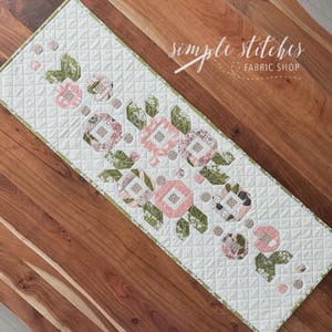 Könnte beinhalten: Gesteppter Tischläufer mit floralem Design. Der Läufer zeigt rosa und gemusterte Blumen mit grünen Blättern und einem hellgrünen Rand. Der Hintergrund ist weiß mit einem Rautenmuster. Der Text "simple stitches FABRIC SHOP" befindet sich oben rechts.