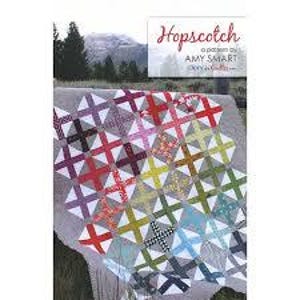 May include: A quilt pattern called "Hopscotch" by Amy Smart. The quilt features a colourful patchwork design with a grid pattern of squares and crosses. The quilt is made with a variety of fabrics in shades of red, orange, yellow, green, blue, purple, and grey.