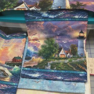 May include: A printed fabric panel featuring a coastal scene with a lighthouse, a house, and a white picket fence under a purple and orange sunset sky. The design includes crashing ocean waves, flying seagulls, and green trees in a landscape.