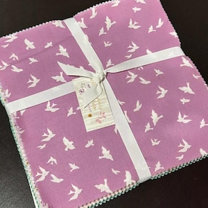 May include: A stack of fabric squares with a white bird pattern on a purple background. The squares are tied together with a white ribbon and a tag that says "Sweetwater" on it.