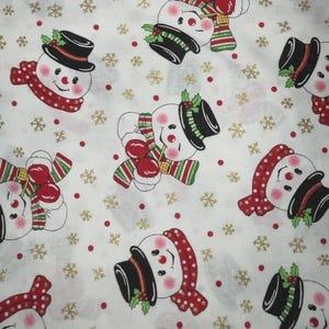 May include: White fabric with a repeating pattern of snowmen, snowflakes, and red dots. The snowmen wear black top hats, red scarves, and mittens. Gold snowflakes are scattered across the background, creating a festive holiday design.