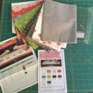 Op de afbeelding: Een set van stoffen vierkanten in verschillende kleuren en patronen, waaronder rood, groen, wit en grijs. De stoffen vierkanten zijn gestapeld, met een grijs stoffen vierkant bovenop. Er is ook een doorzichtige plastic zak en twee gedrukte instructie bladen.