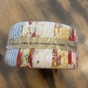 Op de afbeelding: Een rol stof met een patchwork-ontwerp met rode, witte, blauwe en gele bloemenpatronen. De stof is gewikkeld rond een kartonnen buis met een label waarop "www.pennyrosefabrics.com Penny Rose Fabrics" staat.