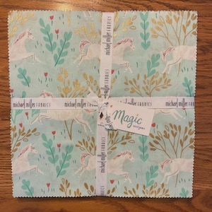 May include: A stack of square fabric pieces featuring a unicorn pattern in white, pink, and gold on a light blue background. The fabric is tied with a ribbon that says "michael miller FABRICS" and a tag that says "Magic sarahjane".