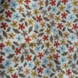 May include: A close-up of a fabric with a repeating floral pattern. The design features small flowers in shades of orange, yellow, and peach, interspersed with brown leaves and small blue star-shaped accents, all set against a white background.