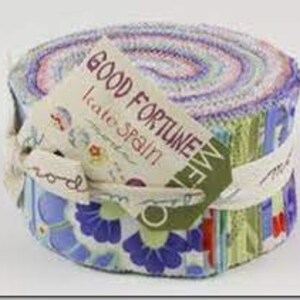 May include: A rolled fabric bundle featuring a mix of floral and patterned fabrics in various colours. The bundle is tied with a white ribbon and a tag that reads "GOOD FORTUNE" and "kate spain". The fabric is likely pre-cut strips, suitable for quilting.