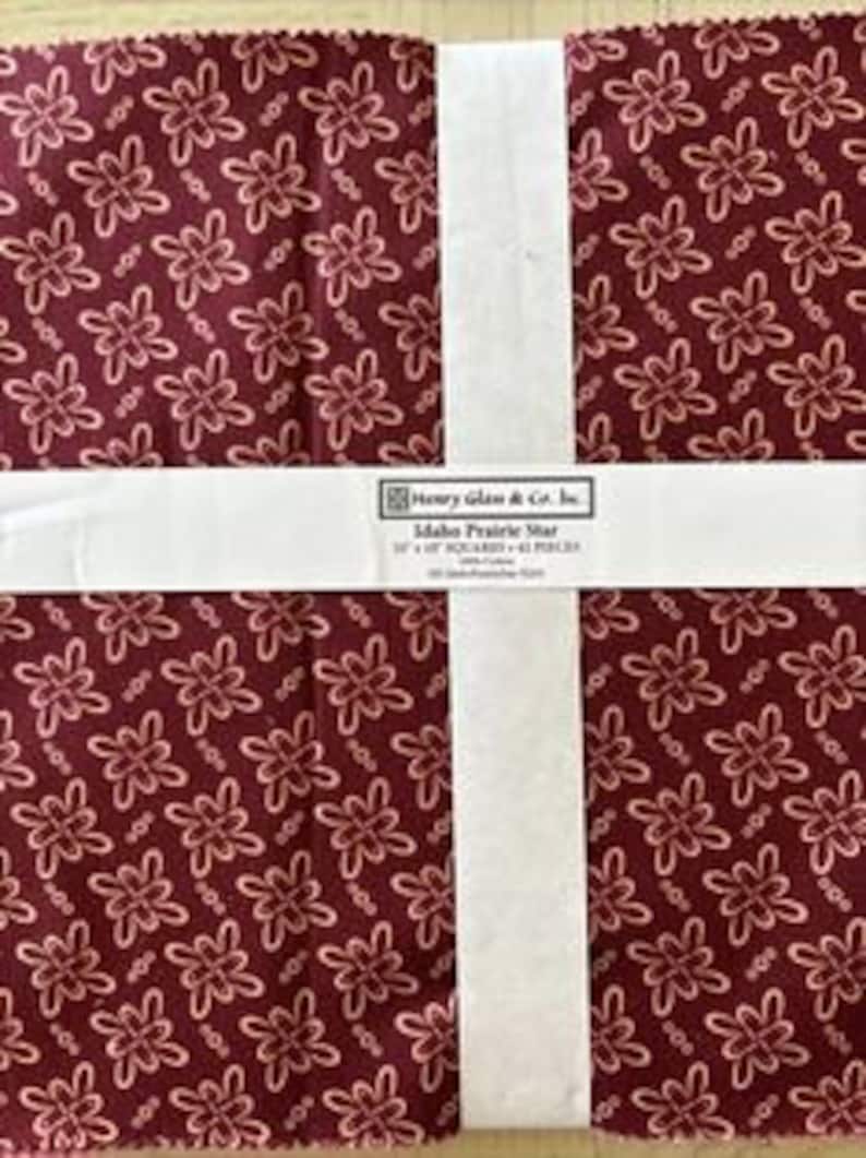 May include: A pack of 12-inch quilt squares featuring a red fabric with a white floral pattern. The fabric is labeled "Idaho Prairie Wheat" by Henry Glass & Co. Inc.