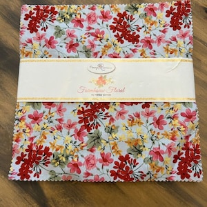 Op de afbeelding: Een pak met stoffen vierkanten met een bloemenprint in tinten rood, roze, geel en groen op een witte achtergrond. De stof is gelabeld "Farmhouse Floral by Nancy Zieman".