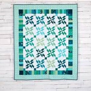 May include: A square quilt with a geometric pattern in shades of teal, green, and white. The quilt features a repeating design of stylized pinwheels, framed by a border of patterned fabric. The quilt is hanging against a white wooden wall.