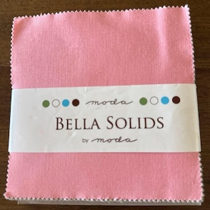 Puede incluir: Un montón de cuadrados de tela rosa con una etiqueta blanca. La etiqueta dice "BELLA SOLIDS" de "moda" con puntos de colores. La tela tiene un aspecto texturizado y un color rosa.