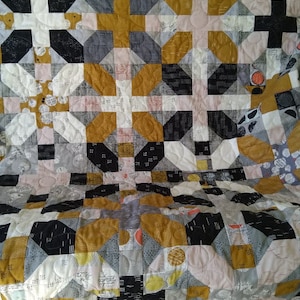 May include: A patchwork quilt featuring a repeating geometric block pattern in mustard yellow, black, white, and light gray. The fabric pieces display various small-scale prints including dots, dashes, and script text, all joined by visible quilting stitches.