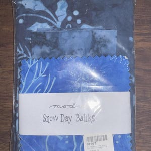 May include: A package of fabric squares in shades of blue, featuring batik patterns. The package includes fabric with a leaf and snowflake design. The label reads "Snow Day Batiks" in a script font.