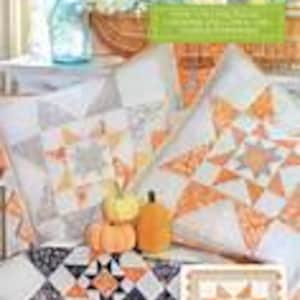 May include: A display of autumn-themed quilted items. Includes two star-patterned throw pillows, a table runner, and a small pumpkin. The pillows and runner feature orange, white, and gray star designs. The text "Stars of the Harvest" is visible.