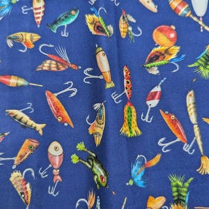 May include: A navy blue fabric featuring a repeating pattern of fishing lures and floats. The lures are in various colours, including red, orange, yellow, green, and blue, with silver hooks. The floats are red and white.