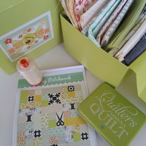 Peut inclure: Une boîte verte avec le texte "Quilter's Gonna Quilt" sur le couvercle. La boîte est ouverte et remplie de chutes de tissu colorées. Un bloc de courtepointe en patchwork de tissu vert et blanc est sur une surface blanche à côté de la boîte.