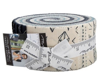 Pode incluir: Um rolo de tecido com fundo branco e desenhos geométricos pretos. O tecido está enrolado e amarrado com uma fita branca. O tecido é etiquetado com "Moda Fabrics - Supplies" e "Moda".