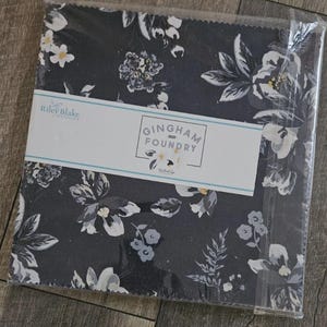May include: A package of fabric squares with a dark floral pattern. The fabric features white, gray, and light blue flowers on a dark background. The label reads "Gingham Foundry 2021" by Riley Blake Designs.