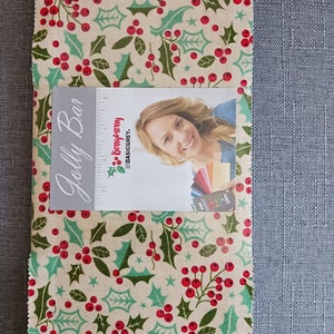 Puede incluir: Un trozo rectangular de tela con un estampado festivo de acebo en rojo, verde y crema. Una tarjeta con el texto "Jolly Bar" y la imagen de una mujer está encima de la tela.