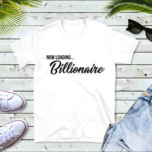 Könnte beinhalten: Weißes T-Shirt mit schwarzem Text, der "NOW LOADING... Billionaire" lautet. Das Shirt liegt flach auf einer Holzoberfläche mit einer Jeans, einer Sonnenbrille und weißen Turnschuhen.