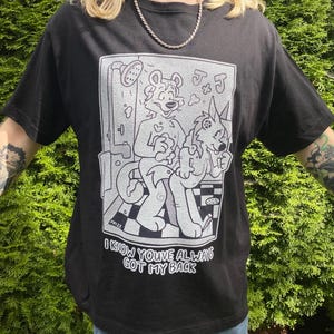 Puede incluir: Camiseta negra con un gráfico de dibujos animados en blanco y negro. El gráfico representa dos animales antropomórficos en una escena de ducha, con el texto "I KNOW YOU'VE ALWAYS GOT MY BACK" debajo. La camiseta tiene mangas cortas.