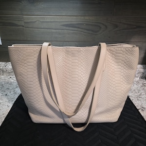 May include: A beige tote bag with a snakeskin pattern. The bag has two long straps and a rectangular shape. The bag is sitting on a black surface.