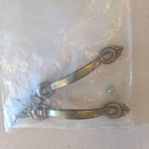 May include: Two curved drawer pulls with an antique brass finish, featuring ornate, detailed mounting plates at each end. The hardware is enclosed in a clear plastic bag, accompanied by small metal screws for installation purposes.