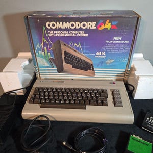 Puede incluir: Un ordenador personal Commodore 64 de color beige con teclado marrón oscuro, acompañado de su caja de cartón original con gráficos coloridos, una fuente de alimentación negra, dos cables de conexión negros y un cartucho HesWare HesModem1.