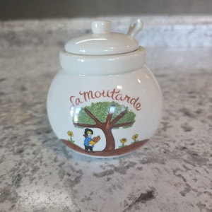 May include: A white ceramic mustard jar with a lid and a small spoon. The jar is decorated with a tree, a person, and the words "La moutarde" in cursive. Yellow flowers are scattered around the base of the tree.
