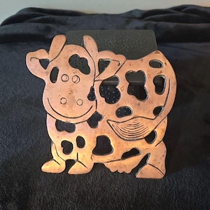 May include: A copper-colored metal trivet shaped like a cow with a cutout design featuring irregular spots, a curved tail, and a smiling face. The surface has a metallic sheen with visible signs of aging and oxidation throughout the metal.