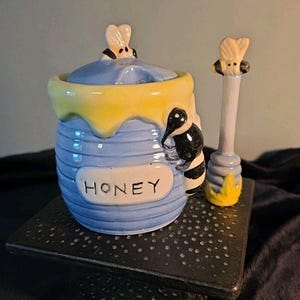 May include: A blue ceramic honey pot with a yellow rim and lid, featuring a bee-shaped handle and a matching spoon. The word "HONEY" is written on the front of the pot. A black and white bee decorates the side of the pot.
