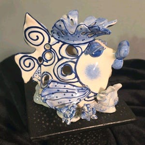 Puede incluir: Una figura de pez de cerámica con base blanca, pintada a mano con patrones de remolinos azules, puntos y orificios circulares recortados. El pez presenta una aleta dorsal prominente, un cuerpo redondeado y elementos decorativos en la base.