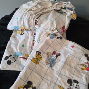 Puede incluir: Una sábana bajera blanca con un patrón de cuadrícula azul claro y un estampado repetitivo de personajes de dibujos animados, incluyendo a Mickey Mouse, Minnie Mouse y Pluto. La tela tiene bordes elásticos y muestra varias poses coloridas.