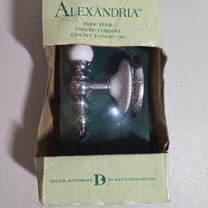 May include: An Alexandria brand robe hook in a chrome and white finish, packaged in a light green cardboard box. The box features the text LIFETIME WARRANTY, ALEXANDRIA, Robe Hook, Gancho Colgador, Crochet à vêtements, and DECOR BATHWARE B BY BATH UNLIMITED.