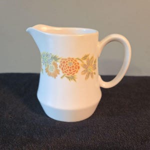 May include: A white ceramic creamer pitcher featuring a horizontal band of floral illustrations in shades of orange, yellow, and blue. The pitcher has a curved handle, a small pouring spout, and a wide base that tapers toward the top rim.